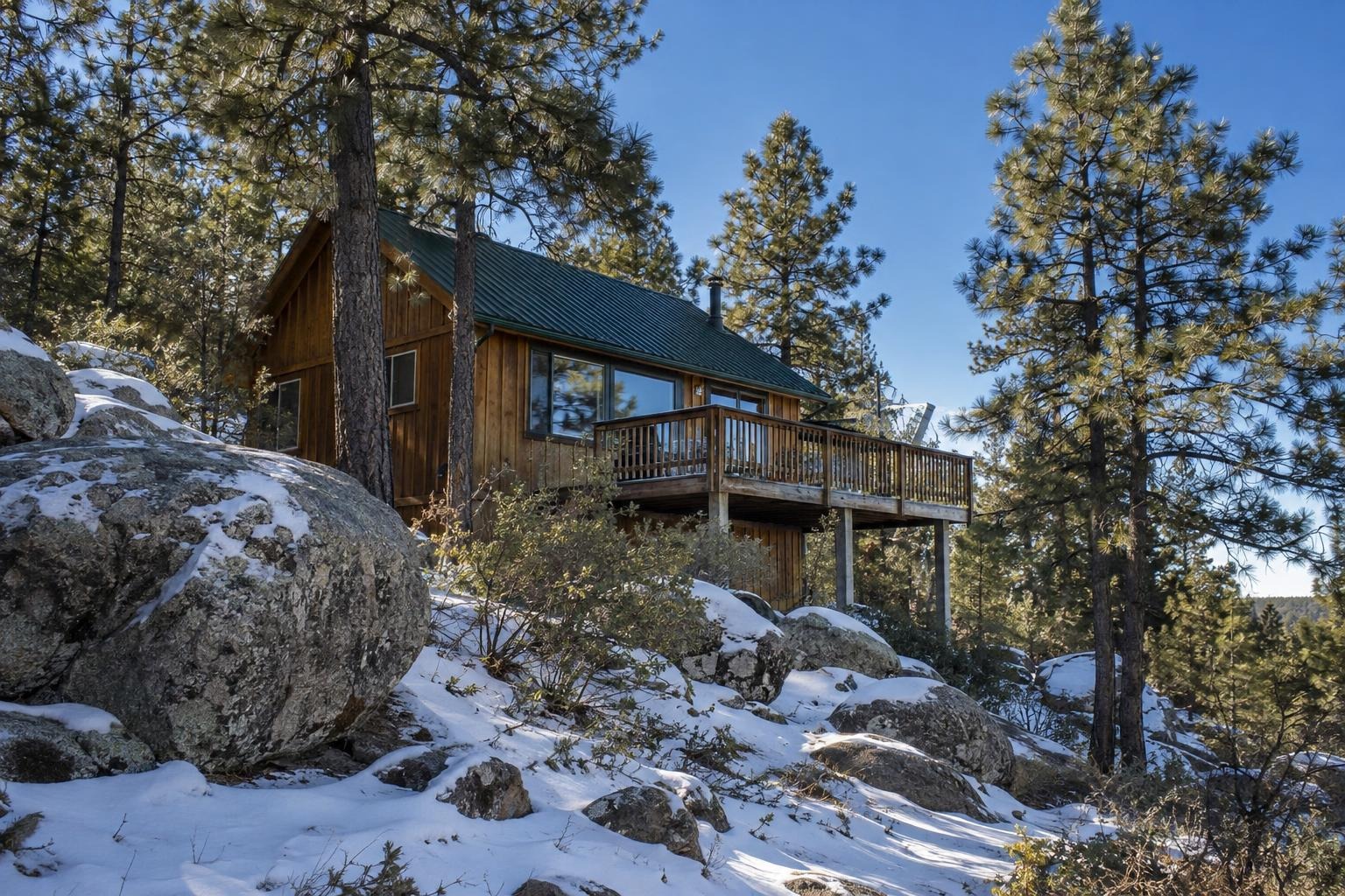 A wood-sided cabin with a green metal roof among pines and snowy granite boulders